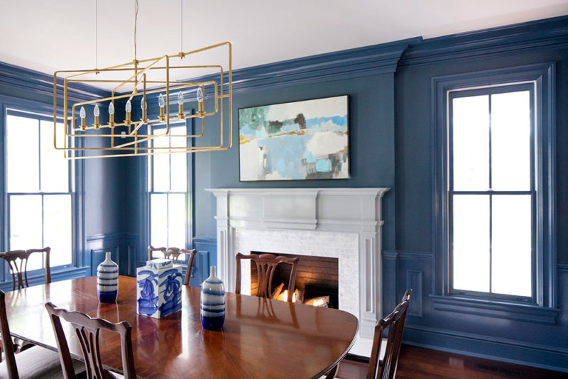 Molly Hirsch Interiors designed this lovely blue dining room with a linear chandelier, marble surround fireplace, and abstract art.